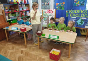 Dzieci siedzą przy stole i słuchają Pani , która tłumaczy zadanie