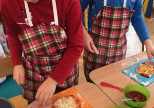Przy stole stoi 2 chłopców przed sobą mają deski na których leży pizza , ozdabiaja swoją pizze