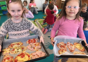 na pierwszym planie stooją 2 dziewczynki, trzymają blachy do pieczenia na których są przygotowane pizze, w tle widać 4 dziewczynki i 2 chłopców siedzących na dywanie
