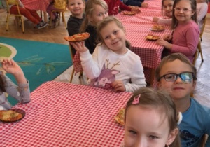 przy pierwszym stole siedzą 2 dziewczynki i chłopiec, przy drugim stole siedzi 6 dziewczynek i 7 chłopców, przy trzecim stole siedzą 2 dziewczynki i 2 chłopców,dzieci degustują pizze
