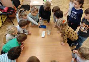 Na stolikach stoją kostkarki do lodu. Dziewczynka nalewa wodę z dzbanka do kostkarek. Wokół stolików stoją dzieci