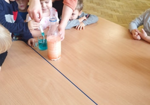Na stole stoi szklanka z pomarańczową wodą, soda, mleko z pomarańczowym barwnikiem. Pani nabiera w strzykawkę niebieska wodą. Dzieci siedzą wokół stołu.