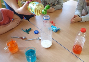 Na stole stoi szklanka z wodą zabarwioną na niebiesko, szklanka z wodą zabarwioną na pomarańczowo, łyżeczki małe. Pani nalewa oleju do mleka. Chłopiec obserwuje.