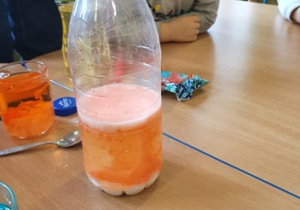 Na stole stoi woda zabarwiona na pomarańczowo, butelka z mlekiem zabarwionym na pomarańczowo. Chłopiec obserwuje butelkę.