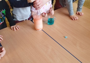 Na stole stoi woda zabarwiona na niebiesko, do butelki z mlekiem zabarwionym na pomarańczowo pani dosypuje sodę.
