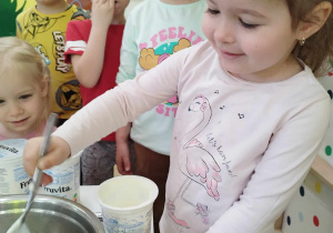 Dzieci oglądają jak dziewczynka przekłada łyżką jogurt do miski.