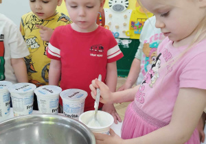 Dzieci oglądają jak dziewczynka przekłada łyżką jogurt do miski.