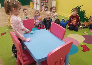 Dzieci oglądają jak dziewczynka układa talerzyki na niebieskim stoliku