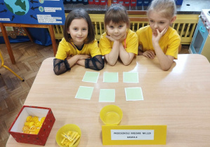 3 dziewczynki w żółtych koszulkach siedzą przy stole i wykonują zadanie