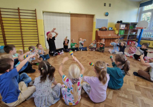 Dzieci siedzą w kole i grają z instruktorem na grzechotkach