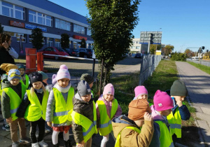 Dzieci w kamizelkach odblaskowych stoją przy urzędzie pocztowym.