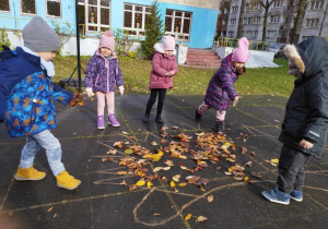 Dzieci wysypują liście na sylwetę jeża narysowanego kredą.