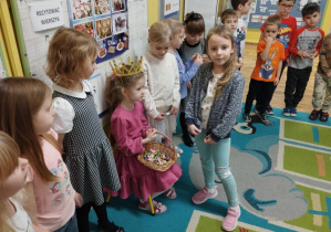 Dzieci stoją w kole na dywanie i składają życzenia koleżance.