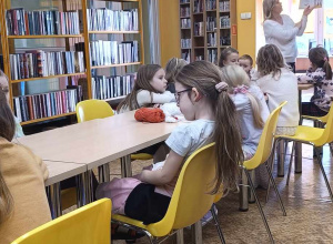 Dzieci siedzą przy stolikach i słuchają historyjki czytanej przez panią bibliotekarkę