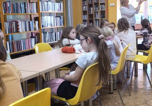 Dzieci siedzą przy stolikach i słuchają historyjki czytanej przez panią bibliotekarkę.