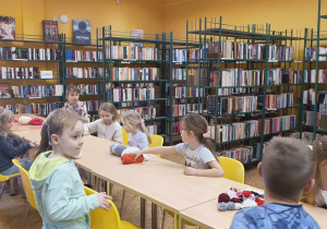 Dzieci siedzą przy stolikach i słuchają historyjki czytanej przez panią bibliotekarkę.