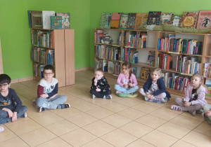 Dzieci siedzą na poduszkach i słuchają opowieści czytanej przez panią bibliotekarkę.