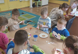 Dzieci siedzą przy stole i wykonują papierowe pączki.