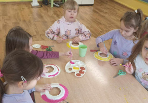 Dzieci siedzą przy stole i wykonują papierowe pączki.