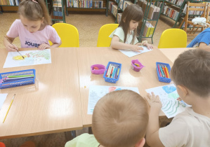 Dzieci siedzą przy stole i kolorują świetliki.