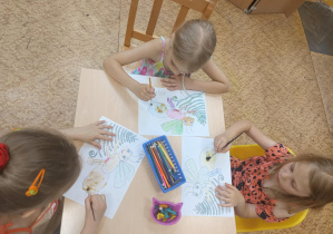 Dzieci siedzą przy stole i kolorują świetliki.