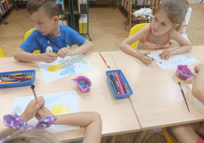 Dzieci siedzą przy stole i kolorują świetliki.
