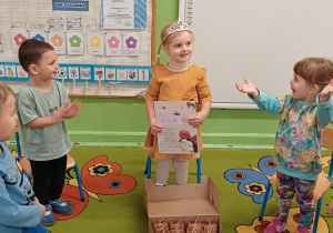 Dziewczynka stoi z dyplomem urodzinowym, dzieci stoją wokół niej