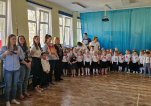 Dzieci oraz dorośli zgromadzeni w sali przygotowują się do odśpiewania hymnu