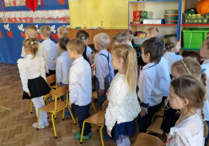 Dzieci oraz dorośli zgromadzeni w sali przygotowują się do odśpiewania hymnu