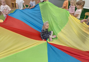 Dziewczynka siedzi w chuście animacyjnej, a dzieci trzymają chustę.