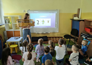 Dzieci siedzą i patrzą na tablicę multimedialną i Panią prowadzącą