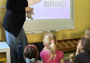 Dzieci siedzą i patrzą na tablicę multimedialną i Panią prowadzącą