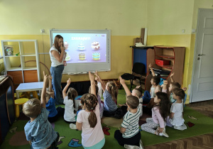 Dzieci siedzą i patrzą na tablicę multimedialną i Panią prowadzącą