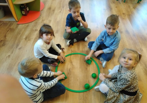 Dzieci siedzą w kole , w środku leży koło w którym są zielone piłeczki i woreczki