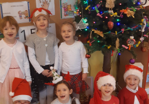 W pierwszym rzędzie siedzą 4 dziewczynki, pierwsza, trzecia i czwarta mają na głowie czapkę mikołajową druga ma opaskę z choinka, w drugim rzędzie stoją 3 dziewczynki,ta w środku ma na głowie świąteczną czapkę