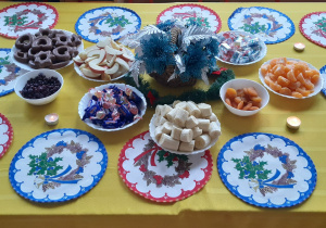N astole leżą: kolorowe serwetki, stroik świąteczny, miski z morelami, żurawiną, banany, jabłka, pierniki, cukierki stół jest przykryty żółtym obrusem
