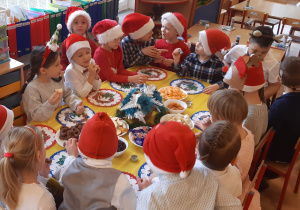 Przy stole siedzą od lewej: dziewczynka, chłopiec, 2 dziewczynki, 6 chłopców, 2 dziewczynki, 3 chłopców, 2 dziewczynki, na stole stoją: cukierki, mandarynki, jabłko, żurawina, banany, stroik świąteczny,kolorowe serwetki, dzieci jedzą smakołyki