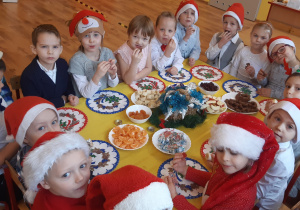 Przy stole siedzą od lewej:3 chłopców, 2 dziewczynki, 3 chłopców, 3 dziewczynki, chłopiec, dziewczynka, 3 chłopców, na stole leżą: bananay, jabłka, morele, żurawina, cukierki cisiastka, stroik świąteczny i kolorowe serwetki