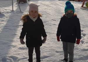 Na pierwszym planie stoją: od lewej dziewczynka i chłopiec, za nimi widać zaśnieżone drzewa i urządzenia terenowe