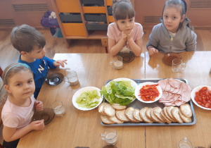 Od lewej strony siedzi dziewczynka, chłopiec i dwie dziewczynki, na środku stołu lerzy taca z kanapkami i warzywami