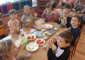 Od lewej strony siedzą 4 dziewczynki, chłopiec, dziewczynka i chłopiec, wszyscy jedzą samodzielnie przygotowane kanapki