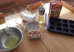 Na stole stoją miska, jajka, mikser, olej, szklanka z łyżką, formemki i opakowanie babeczek