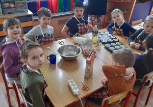 Przy stle siedzą od lewej chłopiec, dziewczynka, chłopiec, chłopiec, dziewczynka, chłopiec, dziewczynka, chłopiec, na stole stoją szklanka, miska, mikser, jajka, foremki i opakowanie babeczek