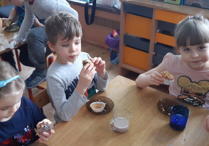 Przy stole siedzą od lewej Dziewczynka chłopiec, dziewczynka, dzieci jedzą mufinki, na stole leżą talerze i kubki