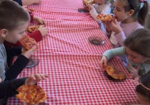 Przy stole siedzą od lewej 5 chłopców, dziewczynka, chłopiec i 3 dziewczynki, dzieci jedzą pizze, na stole leży obrus w kratę