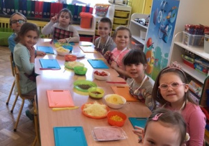Przy stle siedzą od lewej dziewczynka, chłopiec, dziewczynka, chłopiec, 4 dziewczynki, na stole leżą kolrorowe desski, noże, oraz miski ze składnikami do pizzy