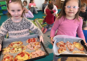 na pierwszym planie stooją 2 dziewczynki, trzymają blachy do pieczenia na których są przygotowane pizze, w tle widać 4 dziewczynki i 2 chłopców siedzących na dywanie