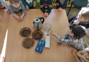 Na stolikach stoją trzy talerze czajnik z ciepłą wodą, czajnik z zimną wodą i dwie kostkarki z lodem. Wokół stolika stoją dzieci