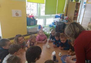 Na stole leży mąka ziemniaczana, chusteczki higieniczne i pianka. Pani robi kule ze sztucznego śniegu, dzieci stoją wokół stołu.