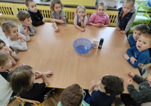 Na stole stoi miska, mąka ziemniaczana i pianka. Dzieci stoją wokół stołu.